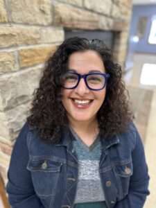 Camp Director Andrea Yenter wears a blue jean jacket, blue striped shirt and black glasses. She has a big smile on her face as she poses for the camera. 